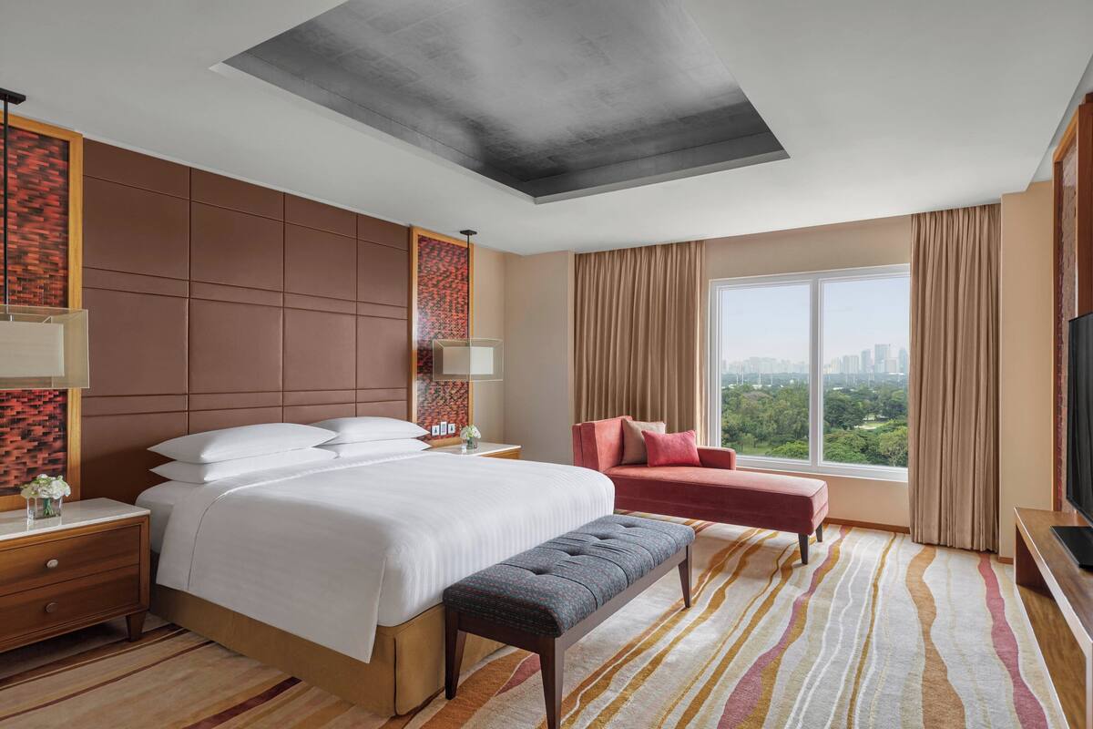 A hotel room with a large bed, a bench, a TV, and a city view.