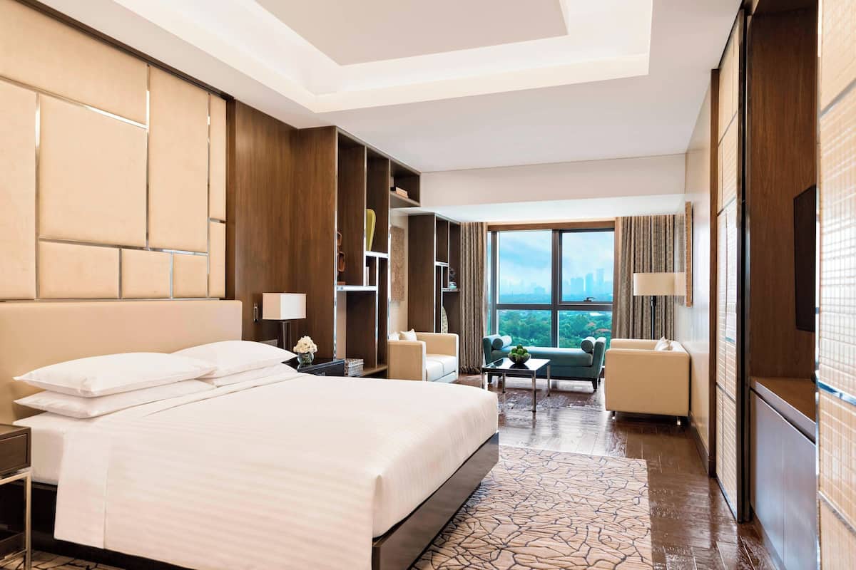 A hotel room with a large bed, a seating area with a sofa and armchairs, a small table, and a large window offering a view of the city.