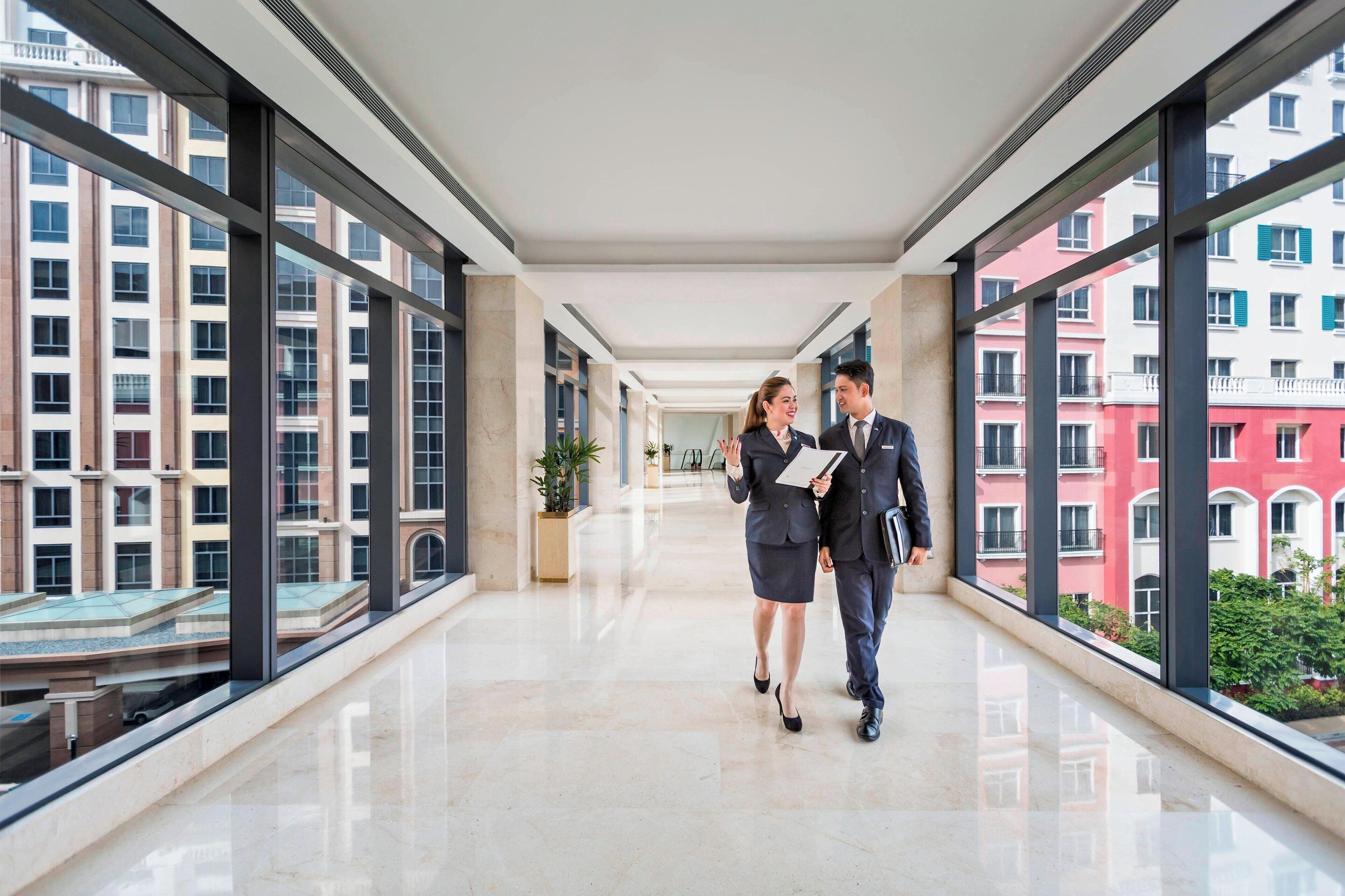 A modern hotel corridor with large windows, a polished floor, and a view of surrounding buildings.