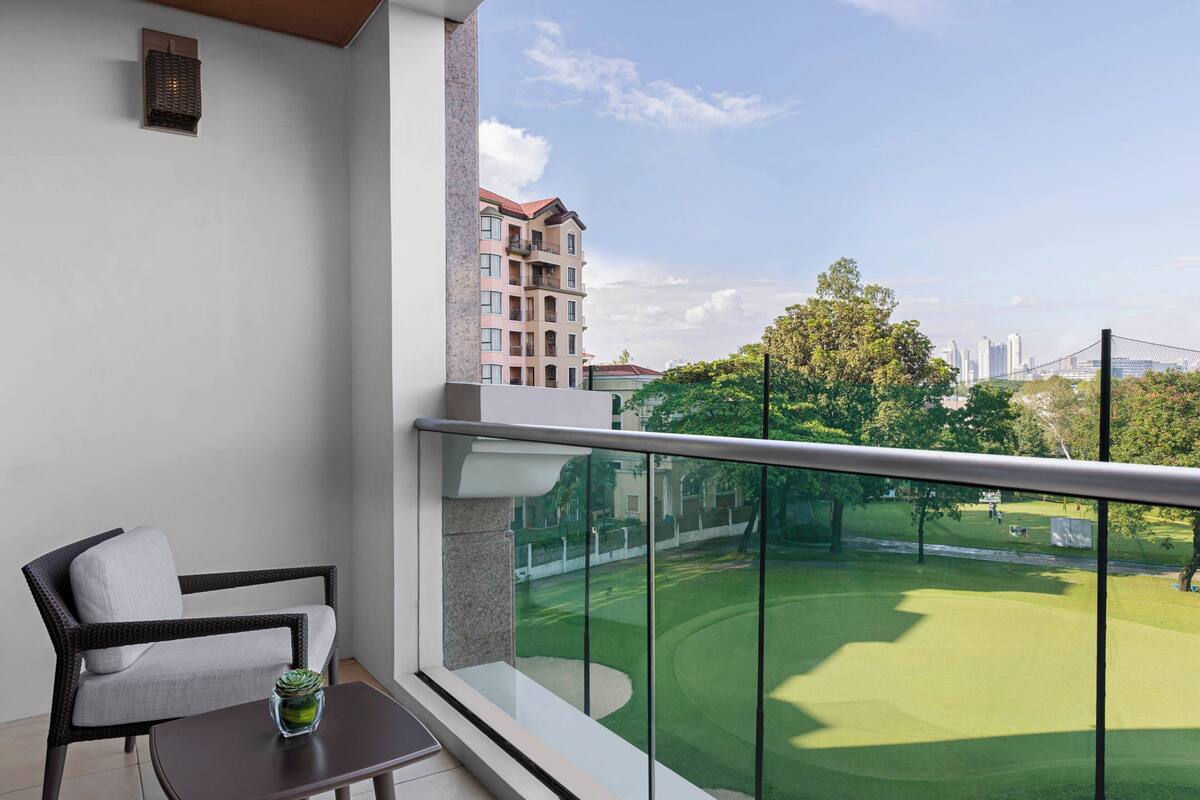 A balcony with a view of a golf course, a chair, and a small table.