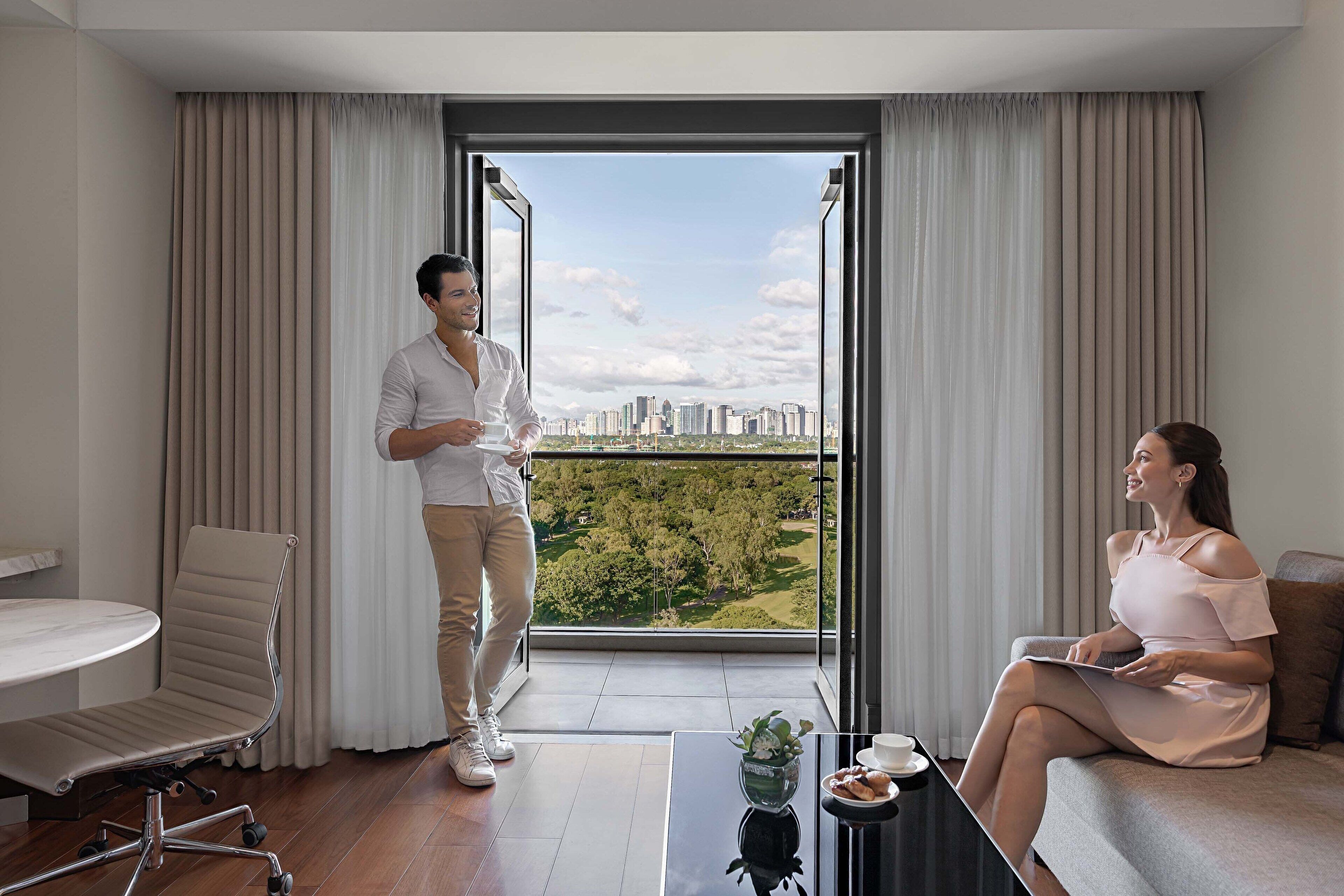 A modern hotel room with a large window offering a city view, a dining table, and a comfortable seating area.