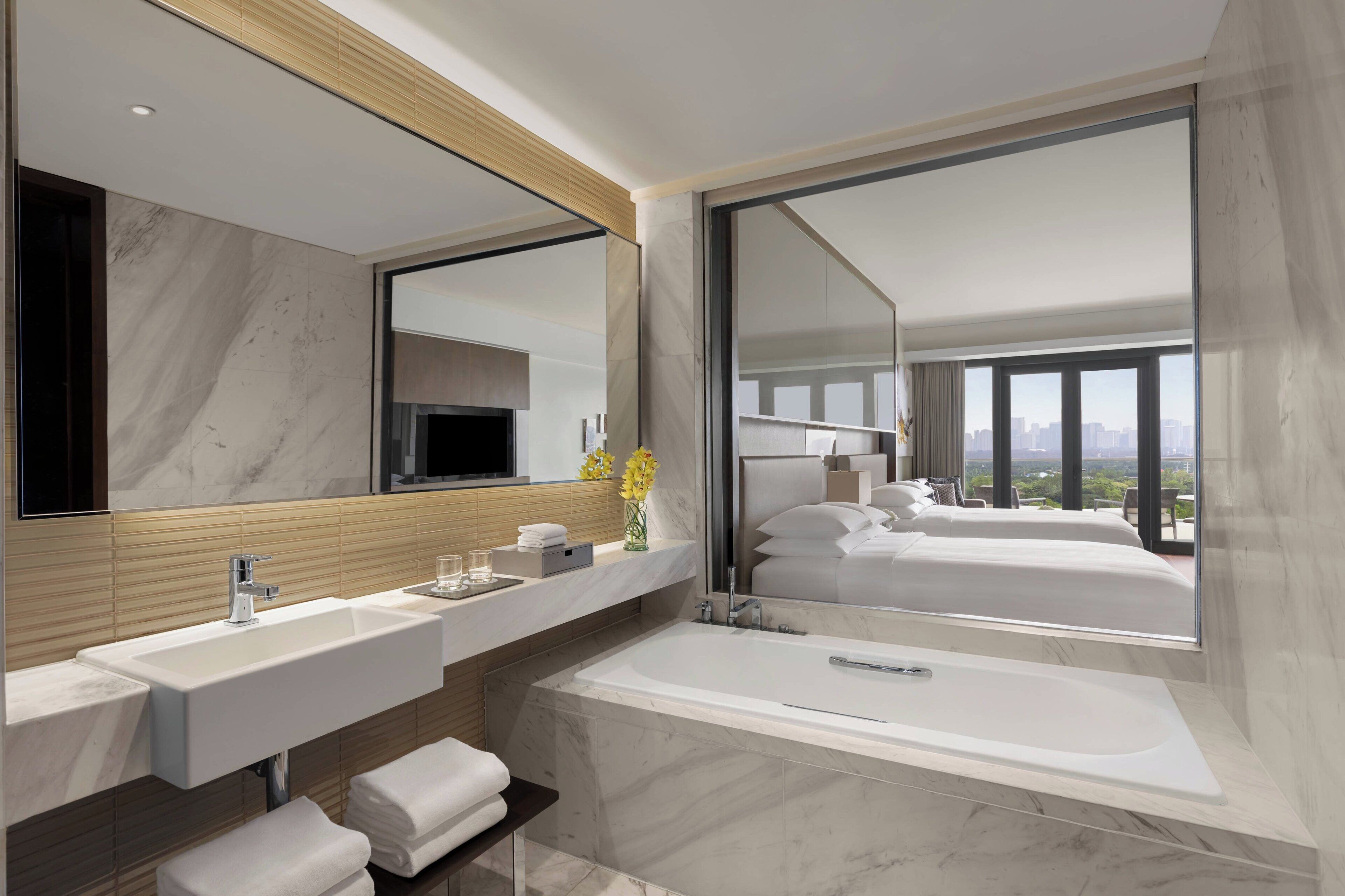 A modern bathroom with a large mirror, two sinks, a bathtub, and a view of a bedroom with a cityscape.