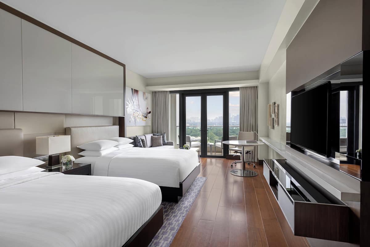 A hotel room with two beds, a TV, a balcony with a city view, and a dining area.