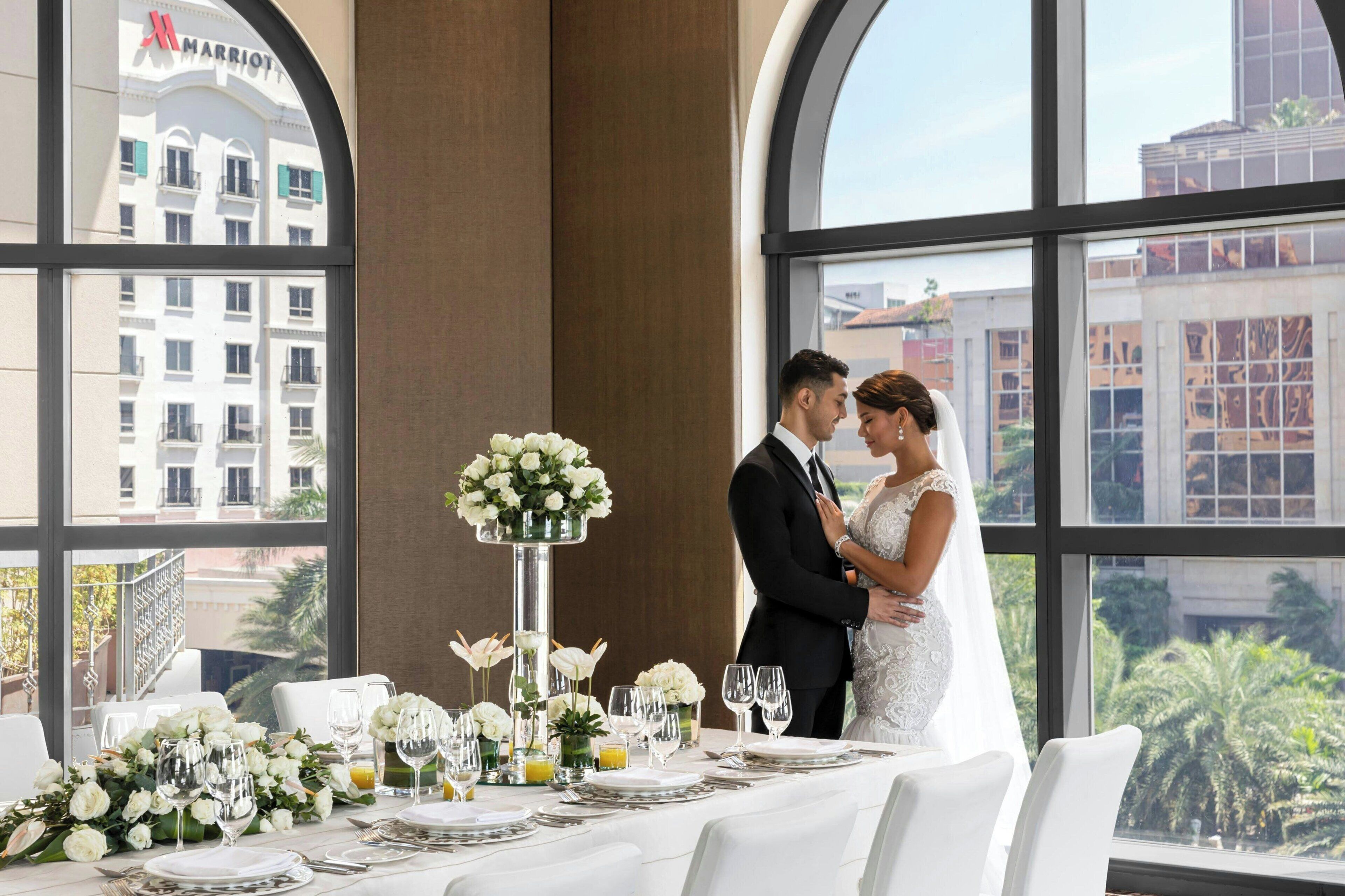A wedding reception table set with white flowers, plates, and glasses, with a view of a building with a 'Marriott' sign.