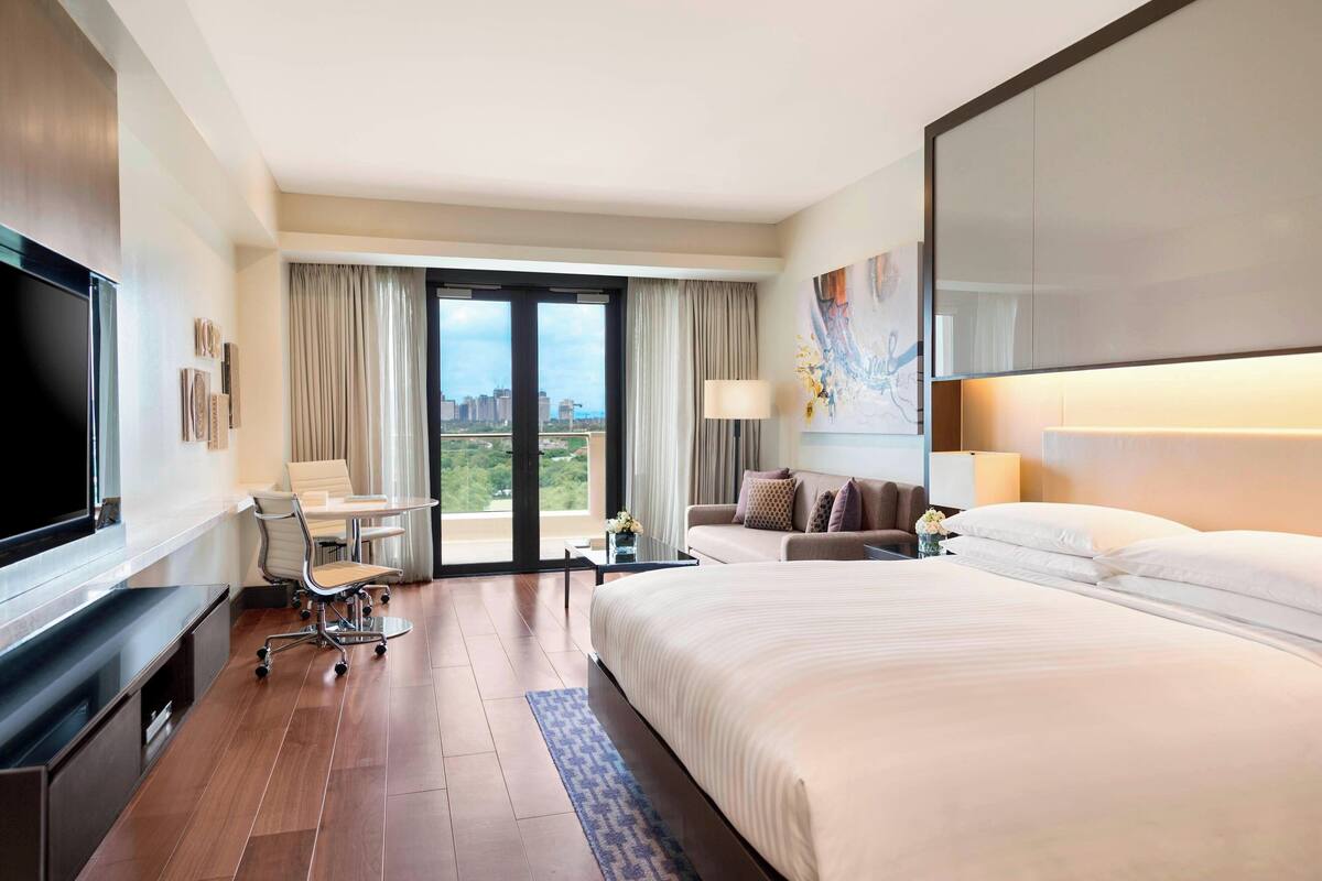 A hotel room with a large bed, a desk with a chair, a television, and a view of the city through the window.
