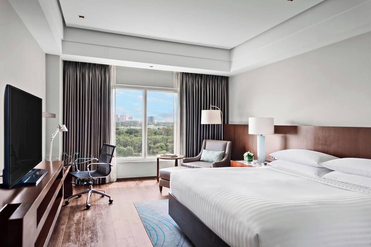 A hotel room with a large bed, a desk with a TV, a chair, a lamp, and a view of the city.