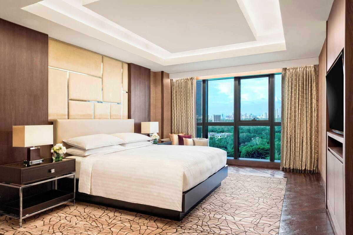 A hotel room with a large bed, a flat-screen TV, a nightstand, and a view of the city through large windows.