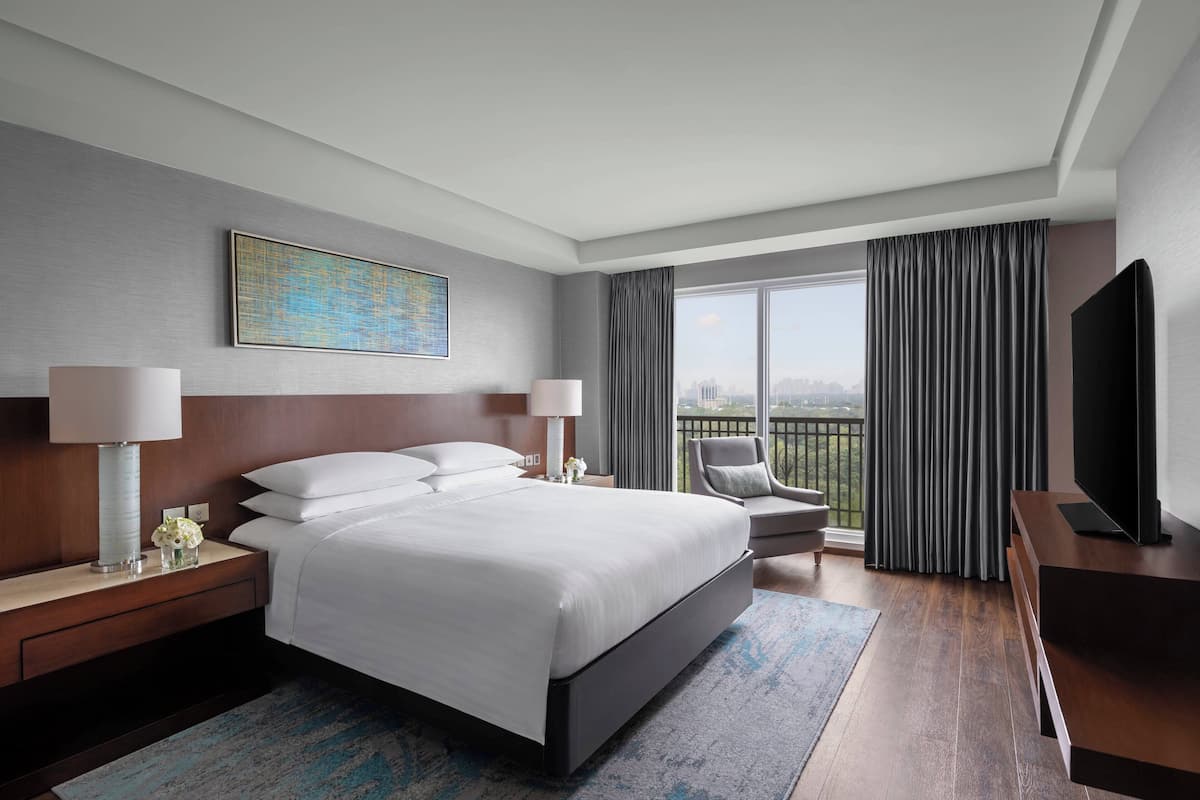 A modern hotel room with a large bed, a desk, a chair, a TV, and a balcony with a city view.