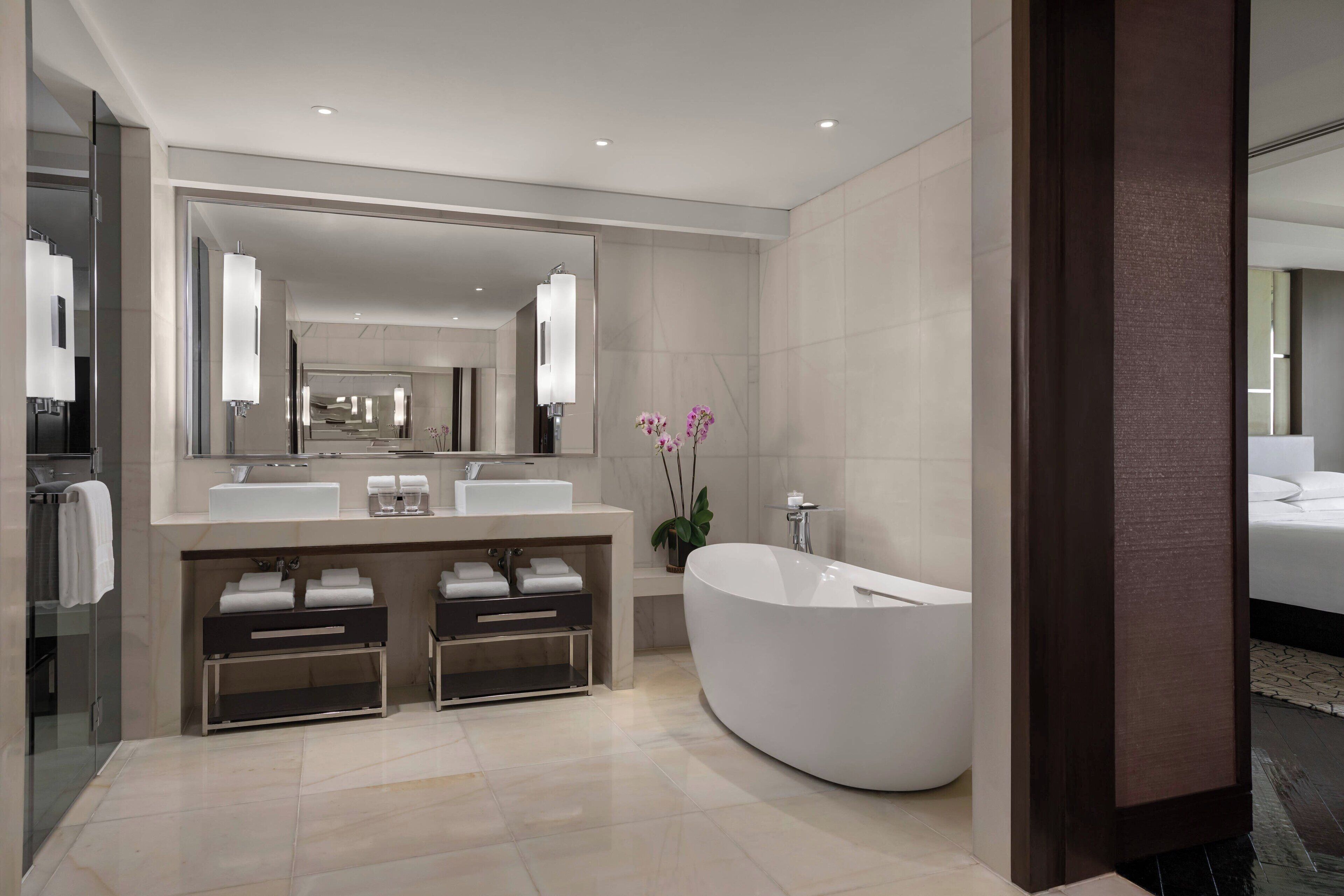 Un baño moderno con un espejo grande, dos lavamanos, una bañera independiente y una orquídea en maceta.