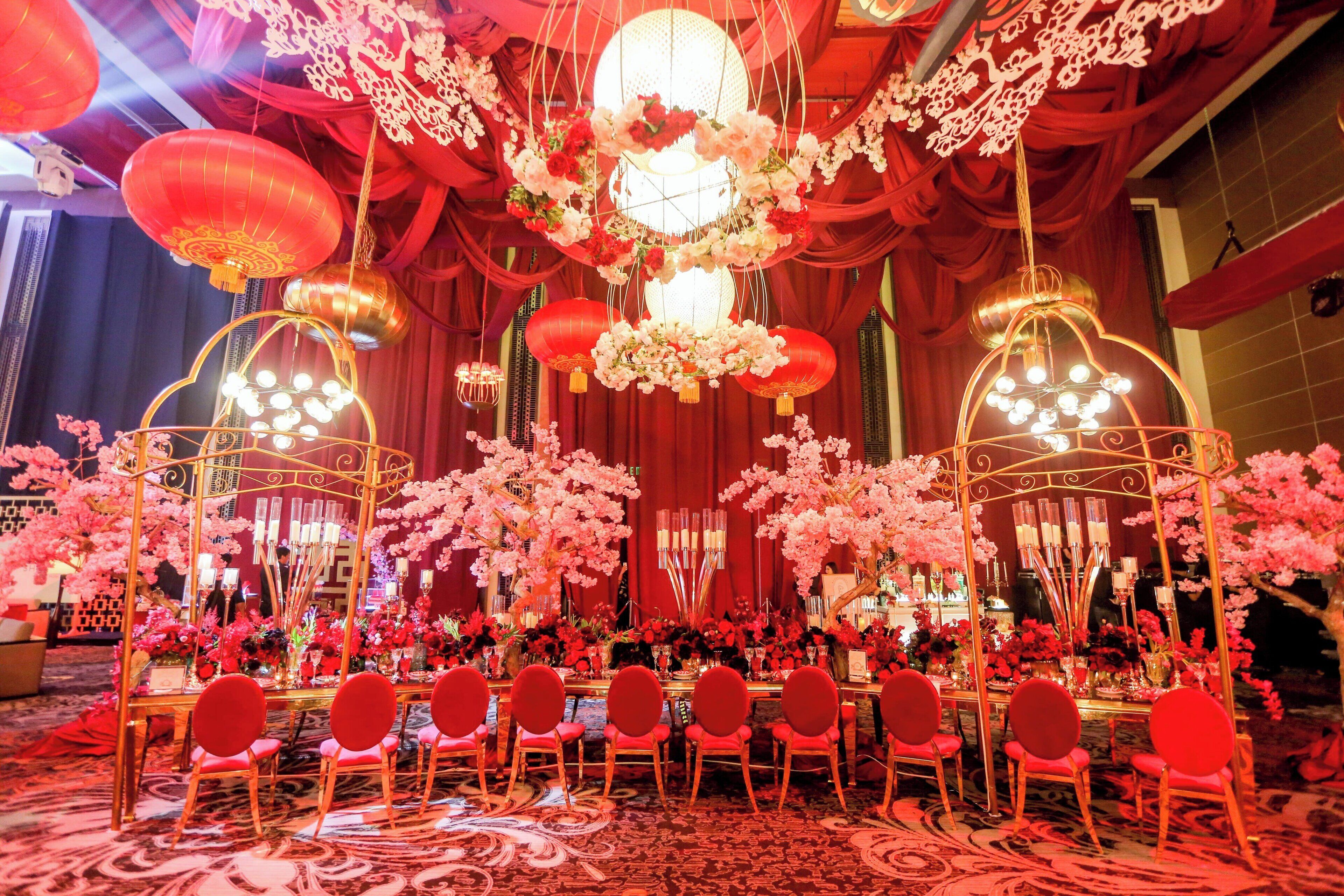 A banquet hall with round tables and red chairs, decorated with floral arrangements and hanging lanterns.