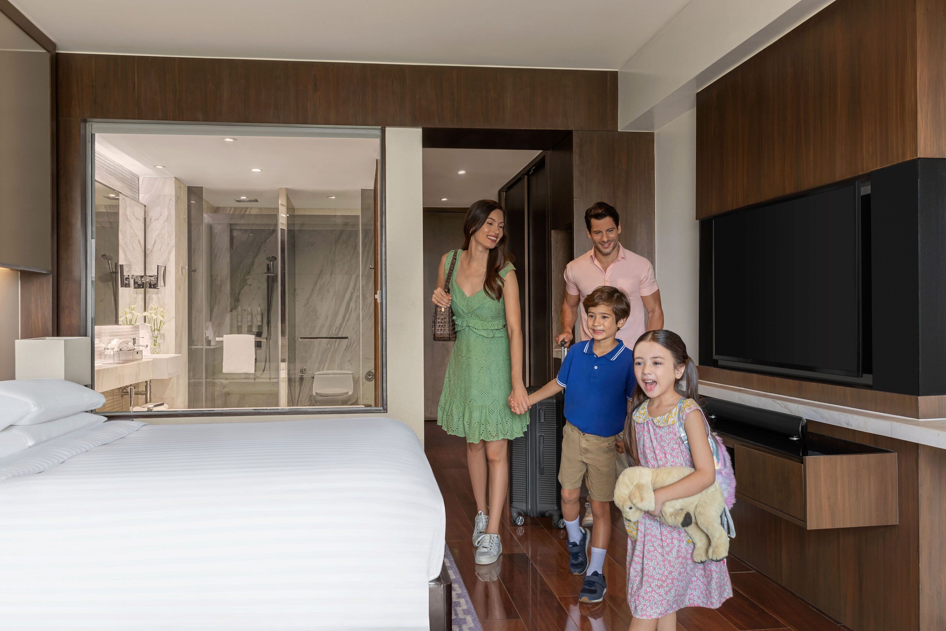 A modern hotel room with a large bed, a glass shower enclosure, a flat-screen TV, and wooden paneling.