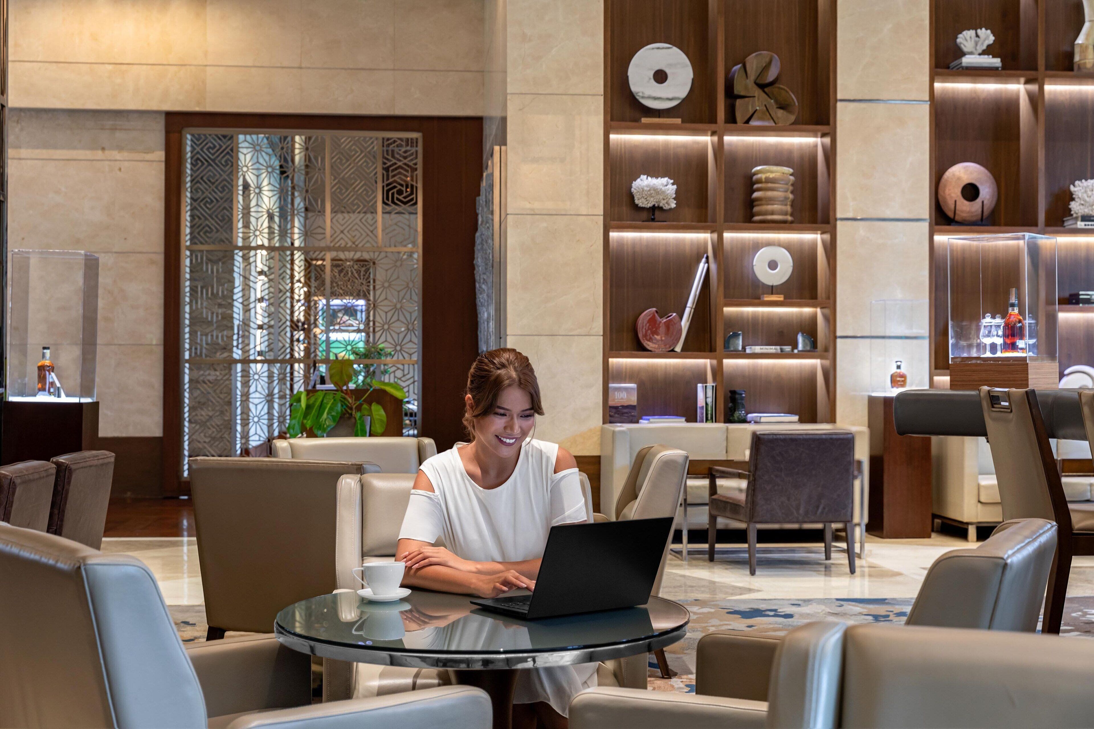 A woman sitting at a round glass table with a laptop, surrounded by leather chairs in a modern lobby with a decorative wall and a reception area in the background.