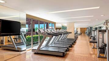 Un gimnasio con varias cintas de correr alineadas en filas, un televisor a la izquierda y diversas máquinas de ejercicio a la derecha.