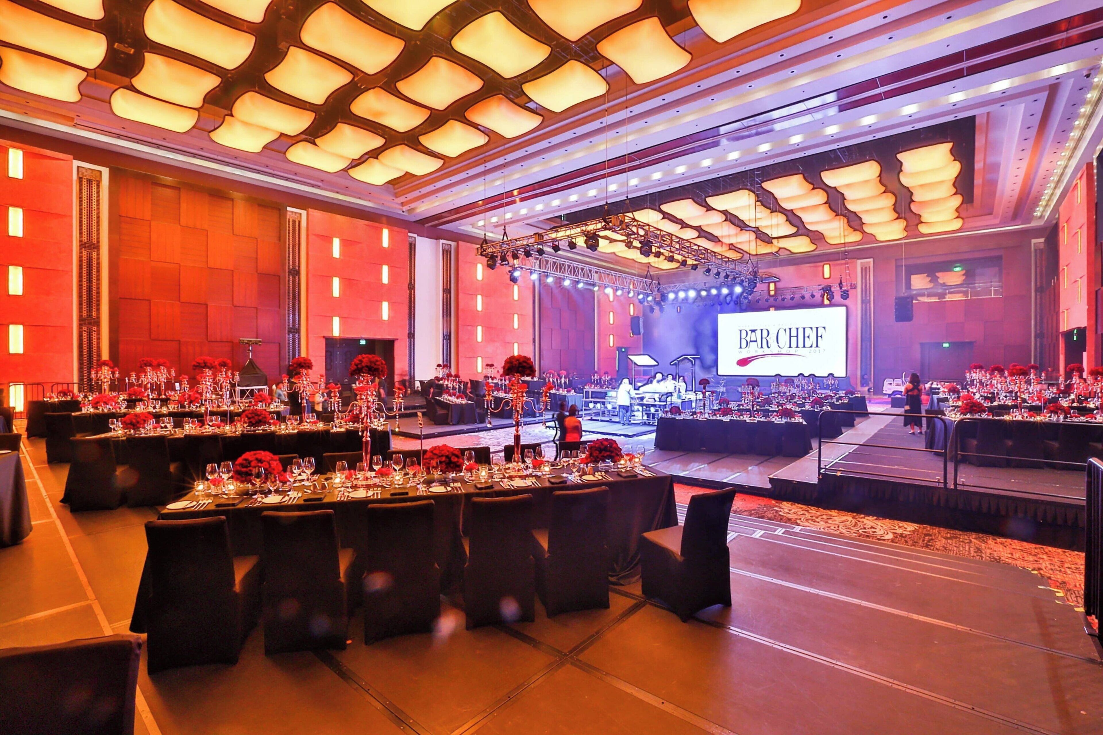 Un salón de banquetes con mesas dispuestas para un evento formal, un escenario con un letrero que dice 'BAR CHEF' y una pantalla grande.
