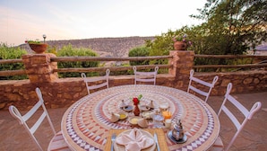 Mesa redonda dispuesta para una comida, con sillas blancas, mantel estampado y vistas a un paisaje montañoso.