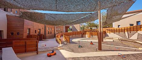 Zona de juegos infantiles con una estructura sombreada, juegos de madera y un pequeño vehículo.