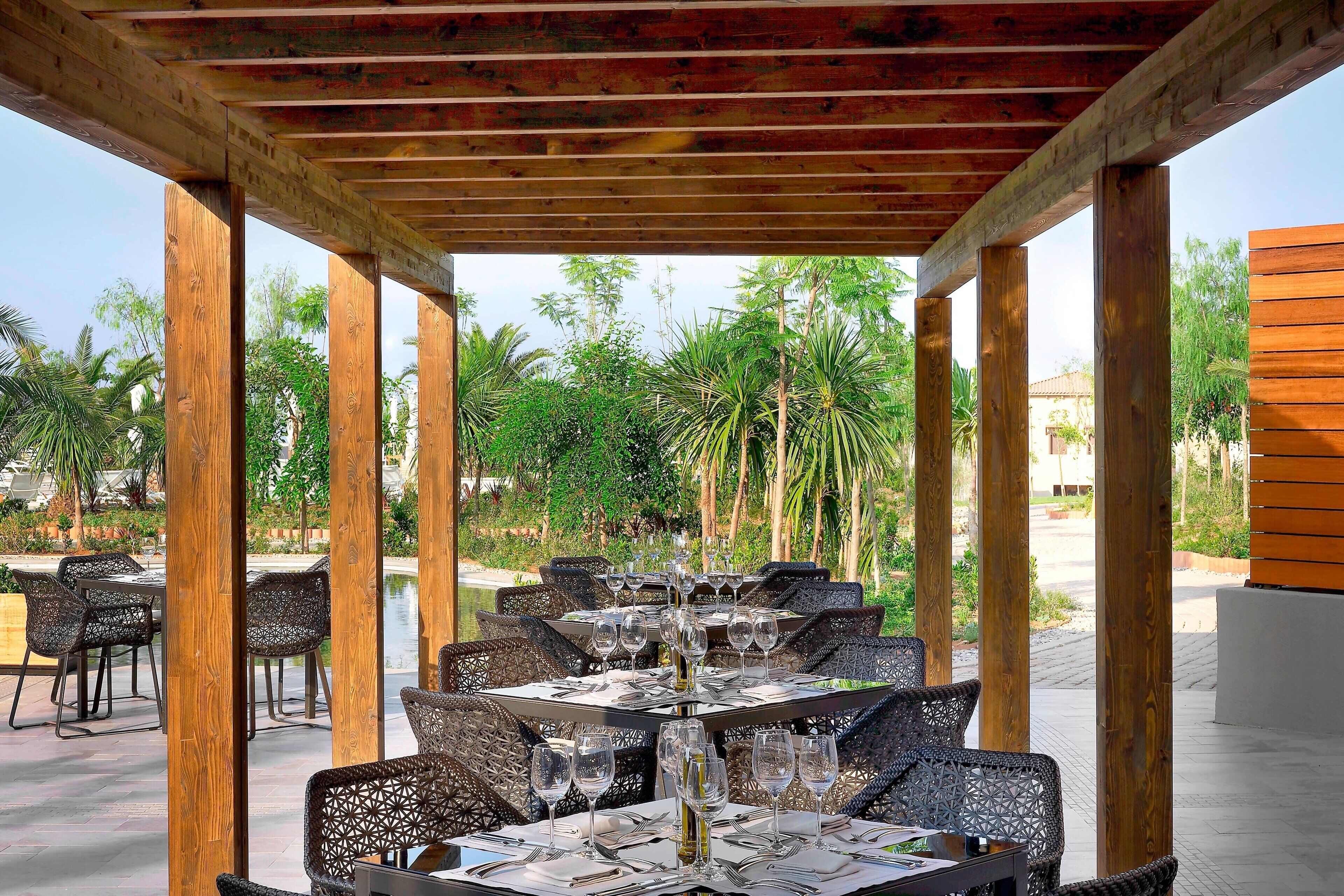 Un área de comedor al aire libre con mesas y sillas, rodeada de pilares de madera y vistas a palmeras.