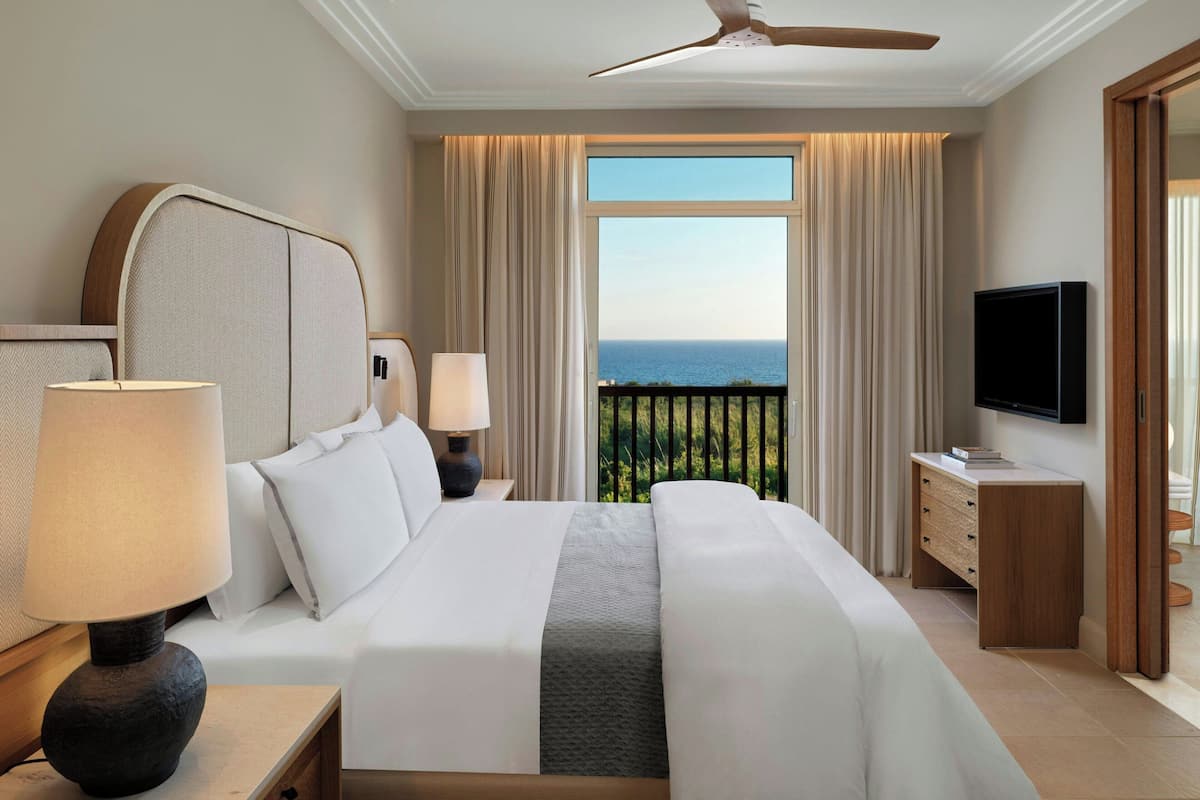 A hotel room with a large bed, two bedside tables, a flat-screen TV, a wooden nightstand, and a view of the ocean.