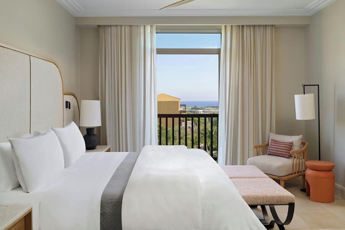 A hotel room with a large bed, a chair, a small table, and a view of the sea.