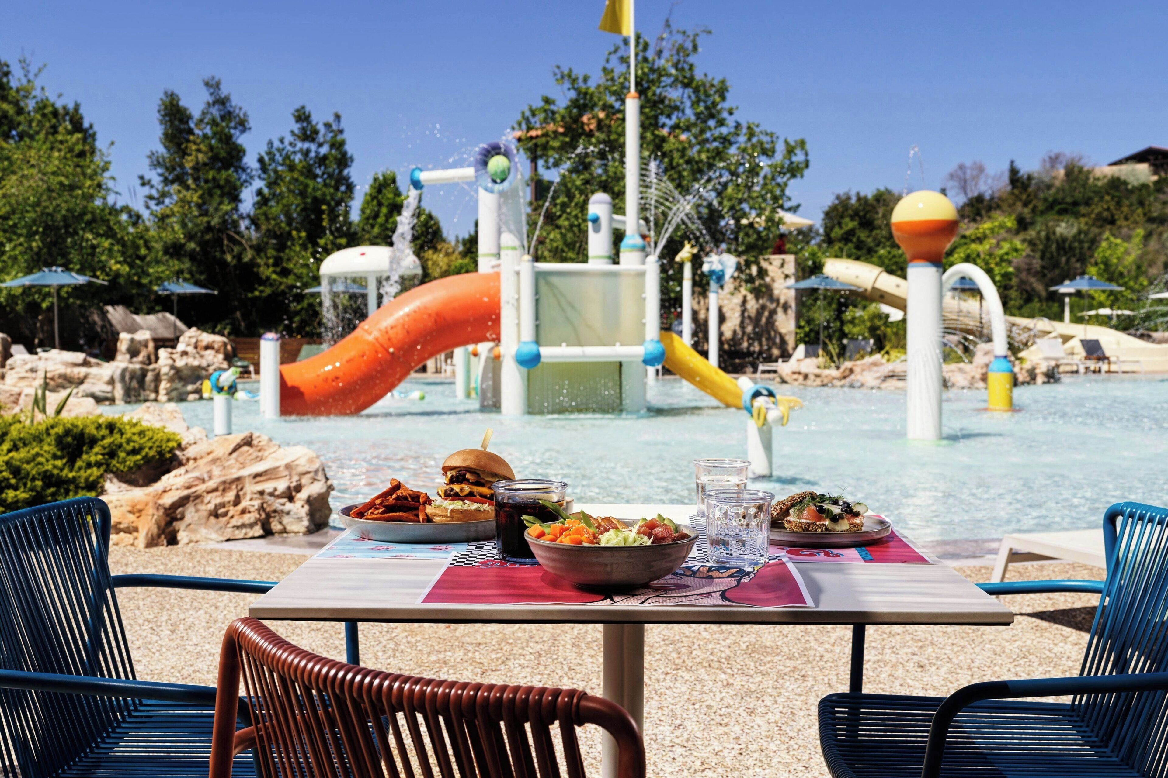 Una mesa de comedor con comida, sillas y un parque acuático al fondo.
