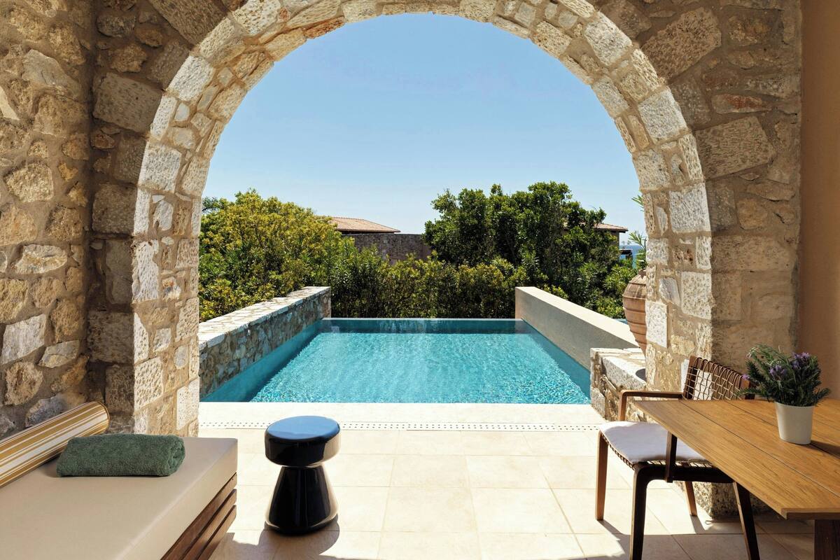 A stone archway leading to a swimming pool with a view of greenery and a building in the background.