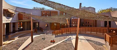 Un patio con un puente de madera, una zona de estar con sombra y un edificio moderno con fachada curva.