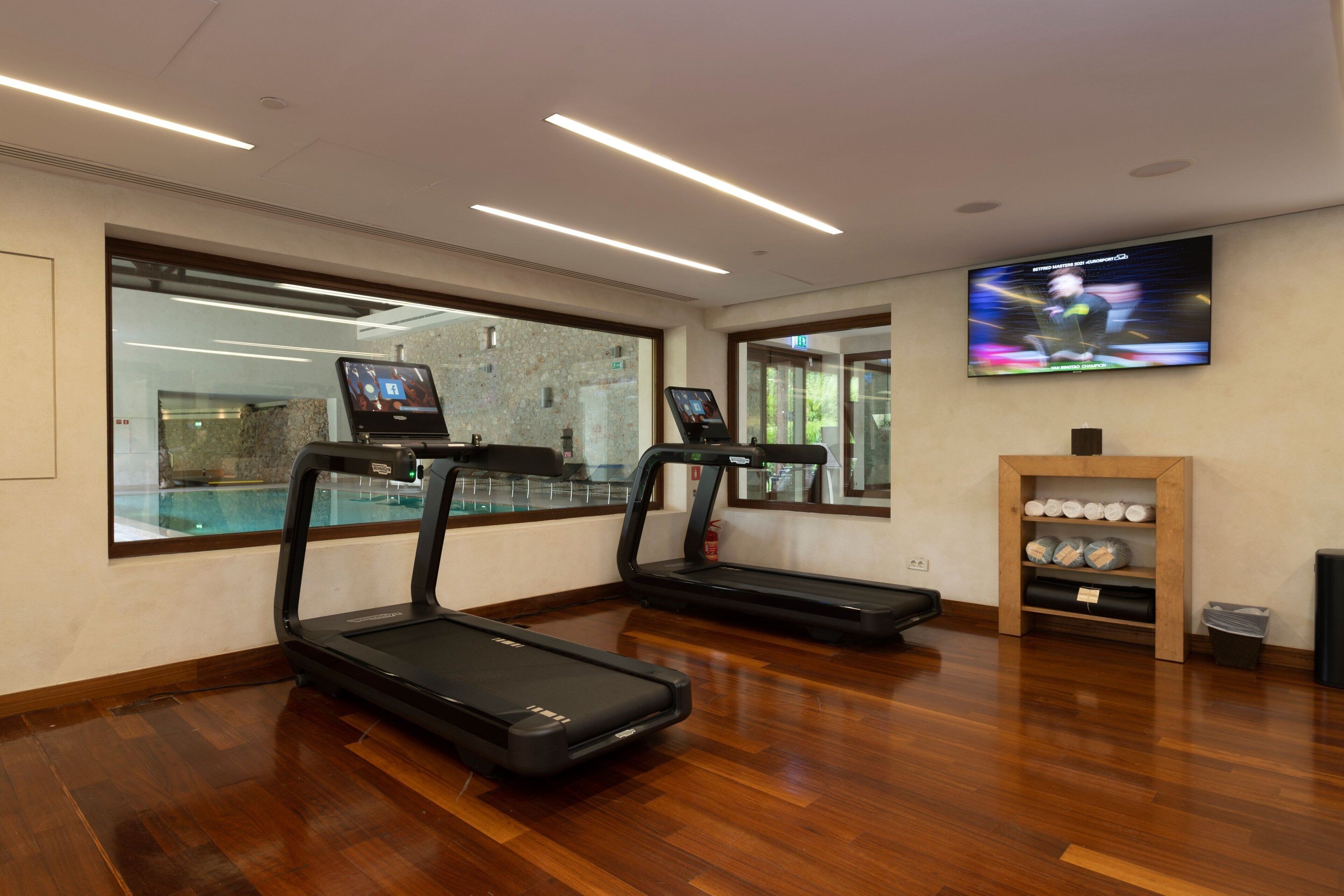 Une salle de sport équipée de deux tapis de course, d’un téléviseur fixé au mur et d’une étagère en bois où sont rangés des accessoires d’exercice.