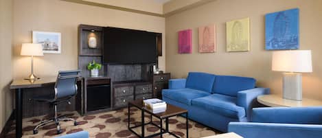 Quarto de hotel com sofá azul, escrivaninha com luminária, televisão de tela plana e obras de arte nas paredes.