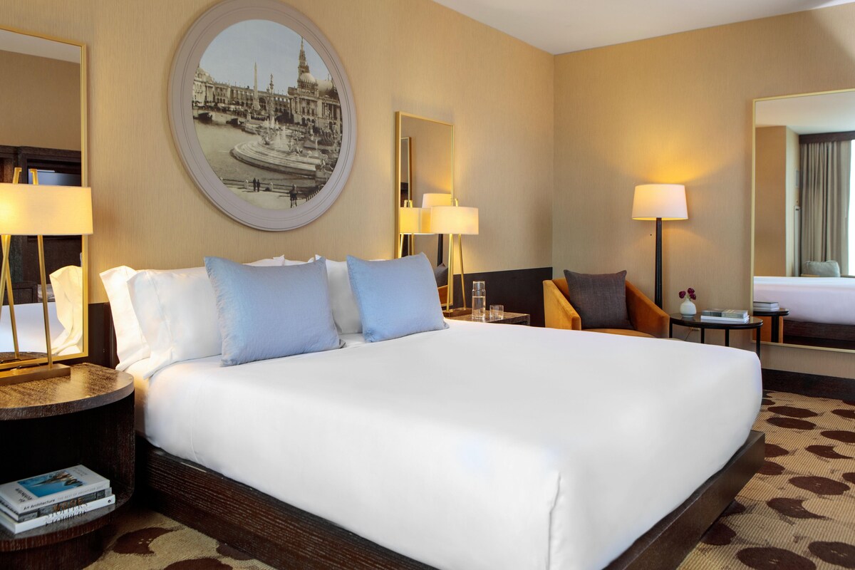 Een hotelkamer met een groot bed, twee nachtkastjes, een ronde spiegel en een wanddecoratie.