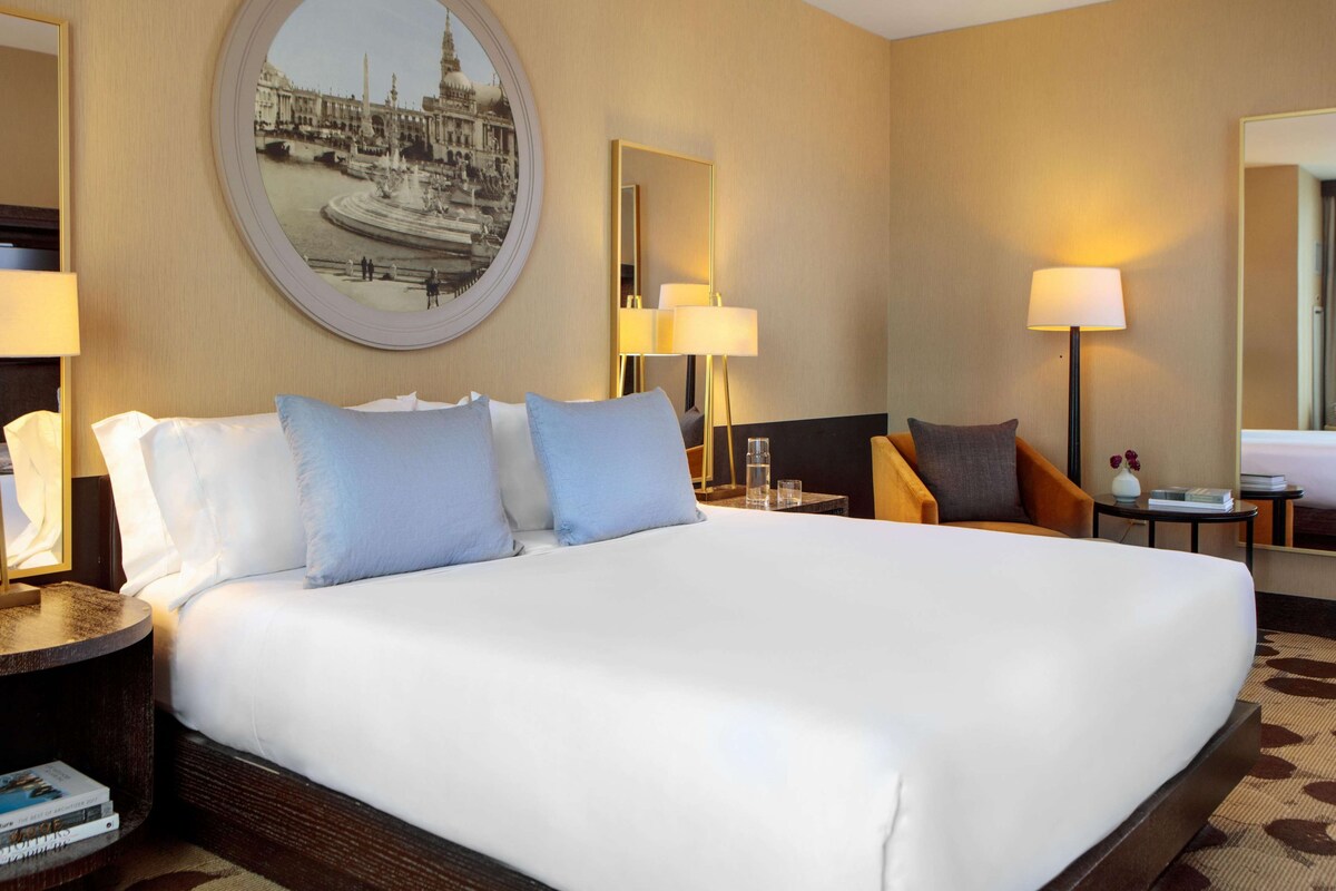 Een hotelkamer met een groot bed, twee nachtkastjes, een ronde spiegel en een wanddecoratie.