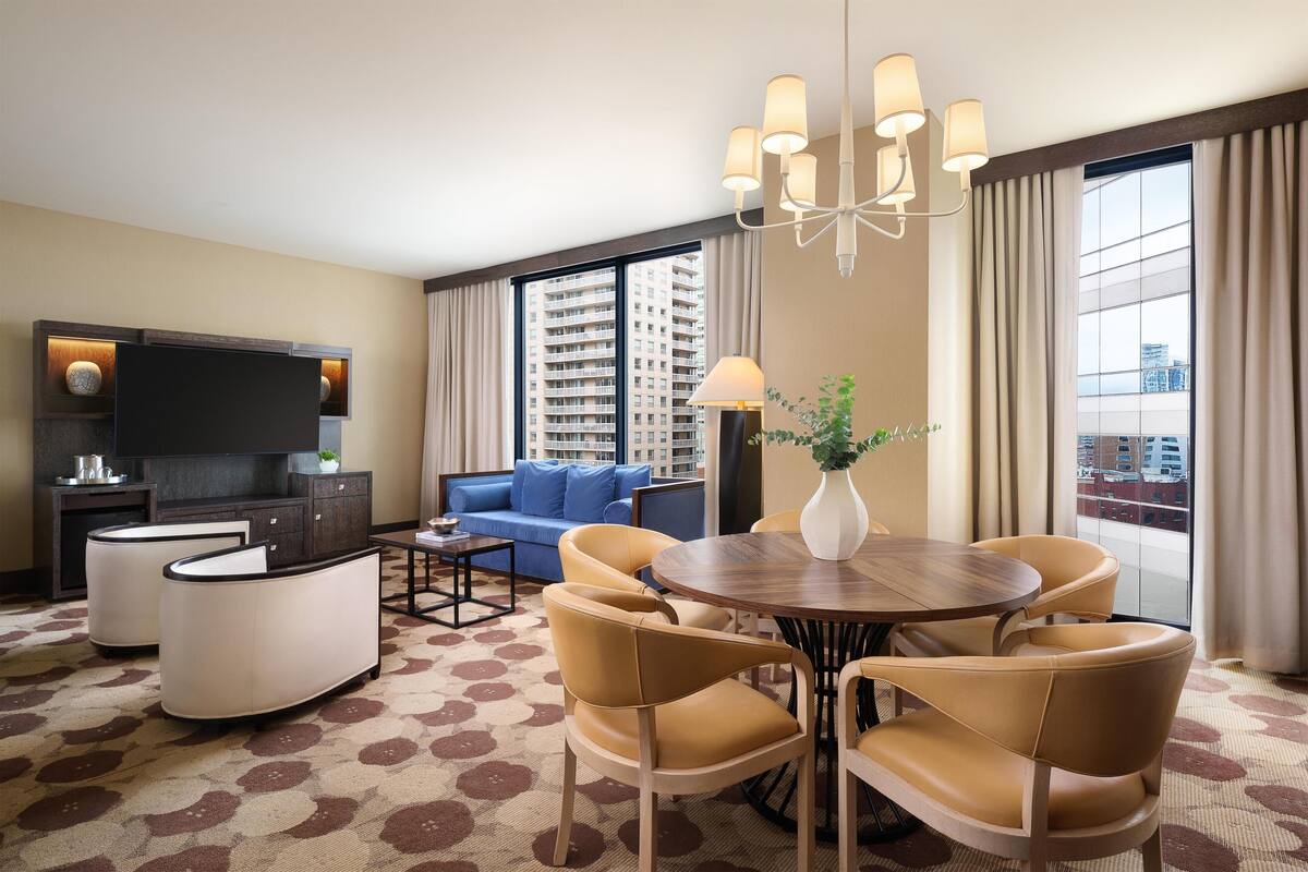 Hotelkamer met een flatscreen televisie, een bank, een eettafel met stoelen en een groot raam met gordijnen.