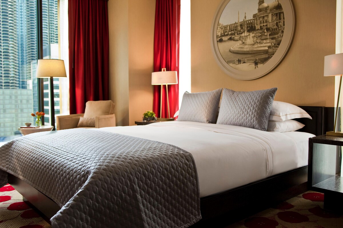 Hotelkamer met een groot bed, rode gordijnen, een ronde wandklok en uitzicht op de stad.
