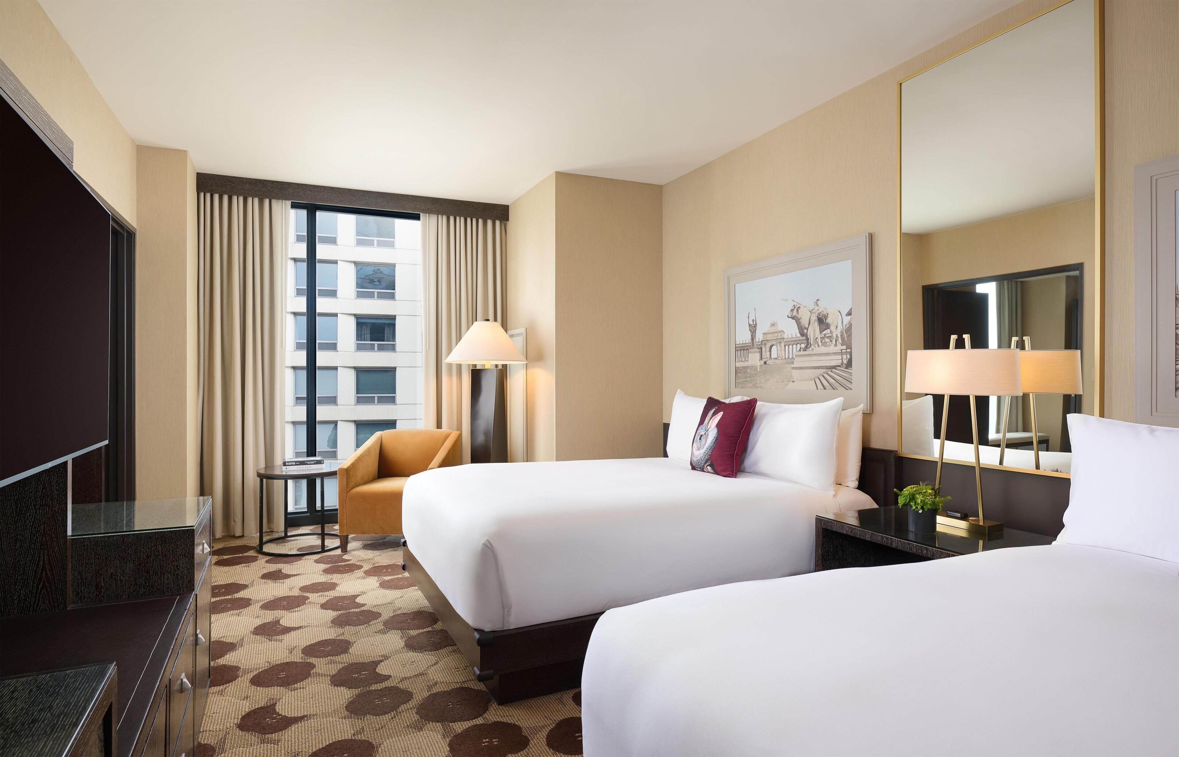 A hotel room with two beds, a desk, a chair, a lamp, and a large mirror.