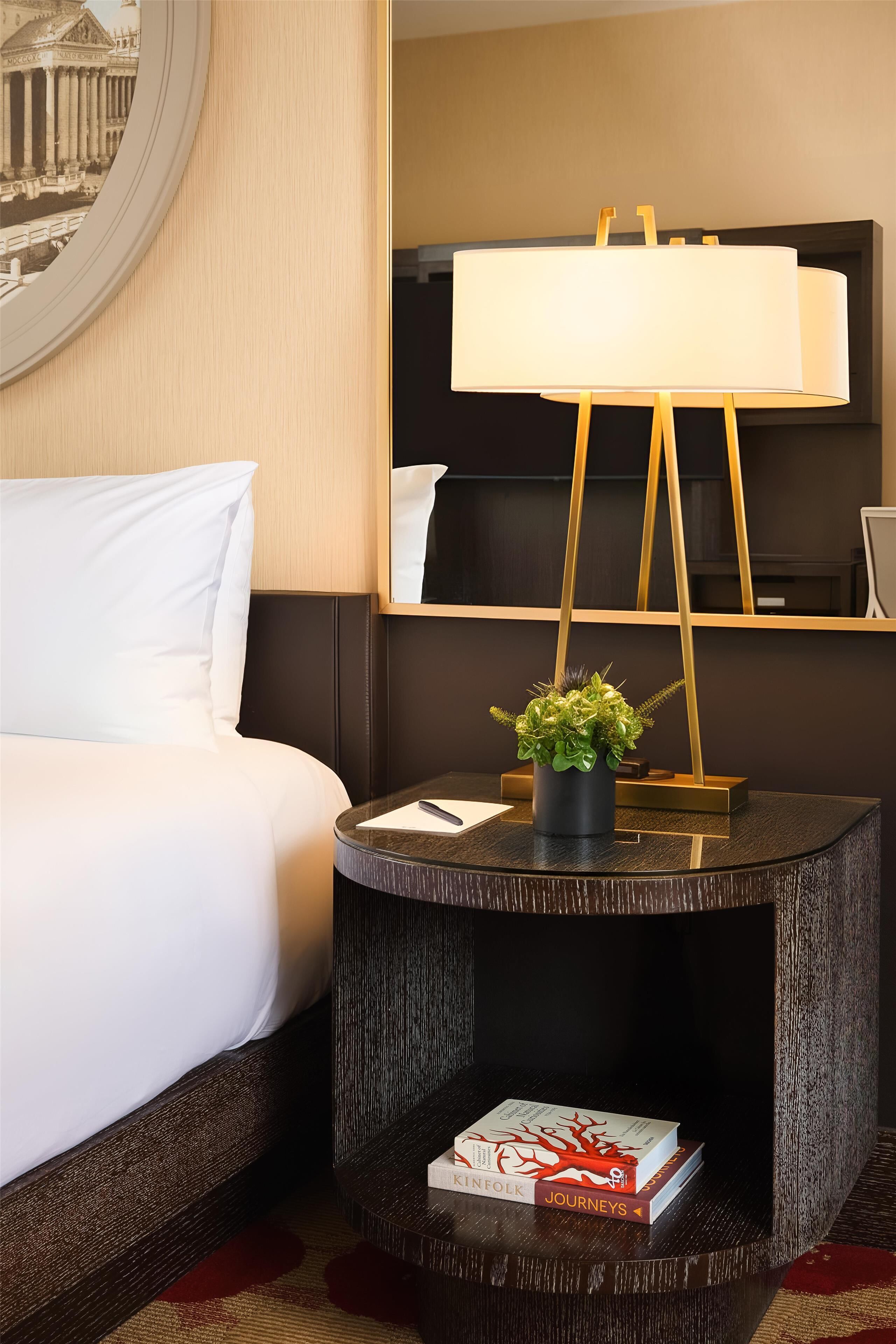 Een hotelkamer met een bed, een rond bijzettafeltje met een lamp, een kleine potplant en een boek getiteld 