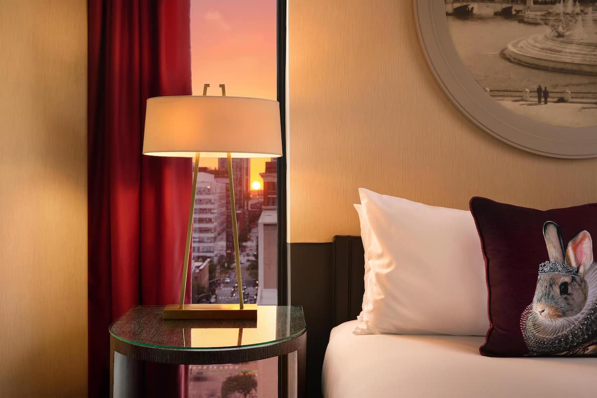 Een hotelkamer met een bed, nachtkastje met een lamp, een rood gordijn en een aan de muur hangend schilderij.