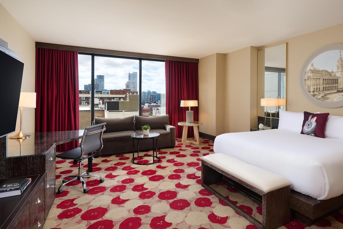 Een hotelkamer met een groot bed, een bank, een bureau en een stoel. Er is een groot raam met uitzicht op de stad.