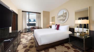 A hotel room with a large bed, a television, a desk with a lamp, and a view of the city through the window.