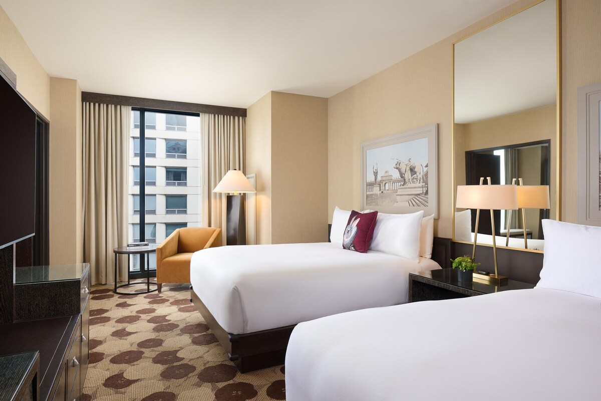Een hotelkamer met twee bedden, een televisie, een klein tafeltje, een stoel en een groot raam met gordijnen.