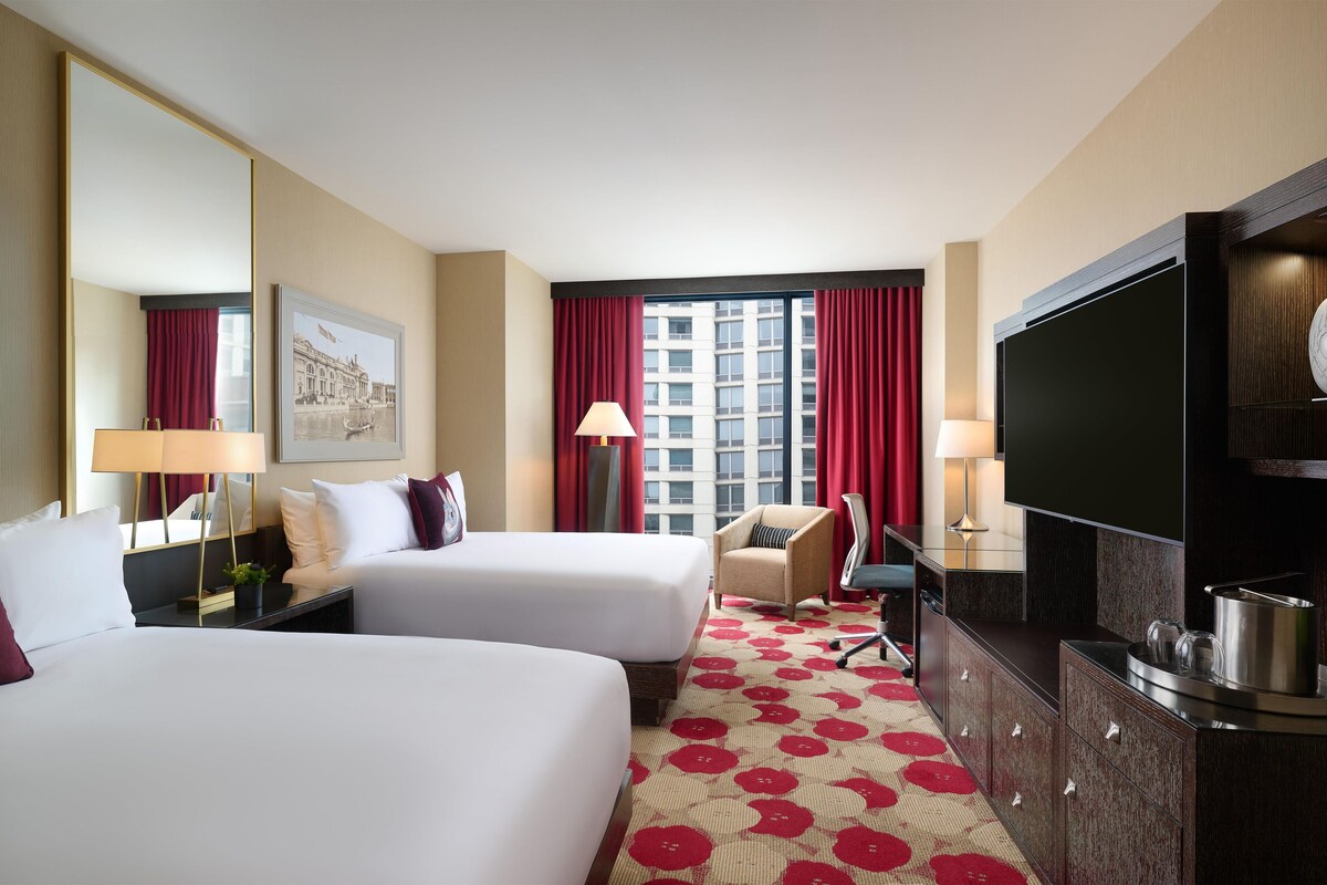 Een hotelkamer met twee bedden, een televisie, een bureau, een stoel en een groot raam met gordijnen.