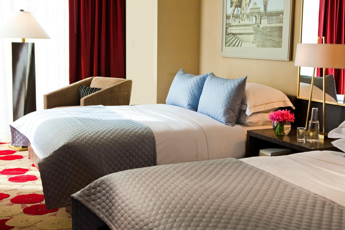Une chambre d’hôtel avec deux lits, une chaise, une table de chevet ornée d’un vase de fleurs et un tableau accroché au mur.