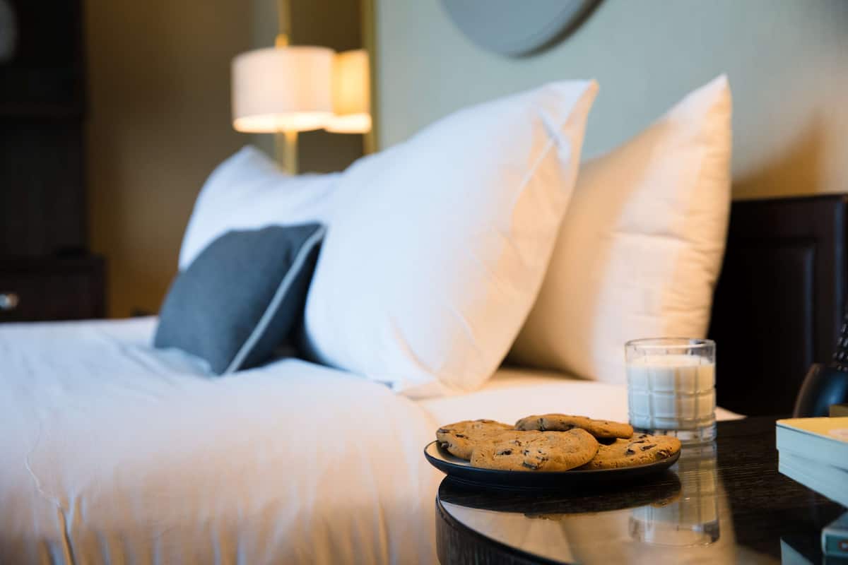 Een net opgemaakt bed met witte en grijze kussens, een bord koekjes en een glas melk op een nachtkastje.