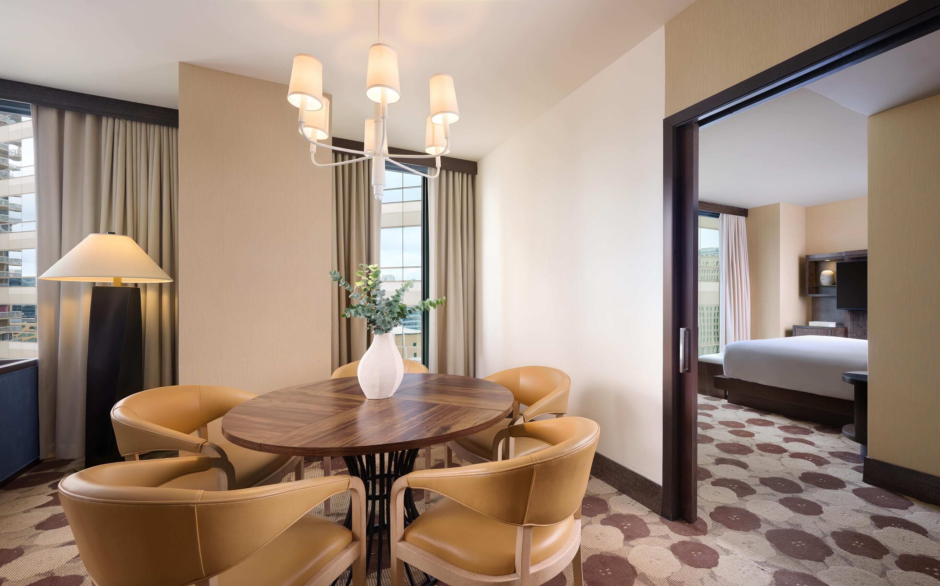A hotel room with a round dining table, six chairs, a vase with greenery, a lamp, and a bed.