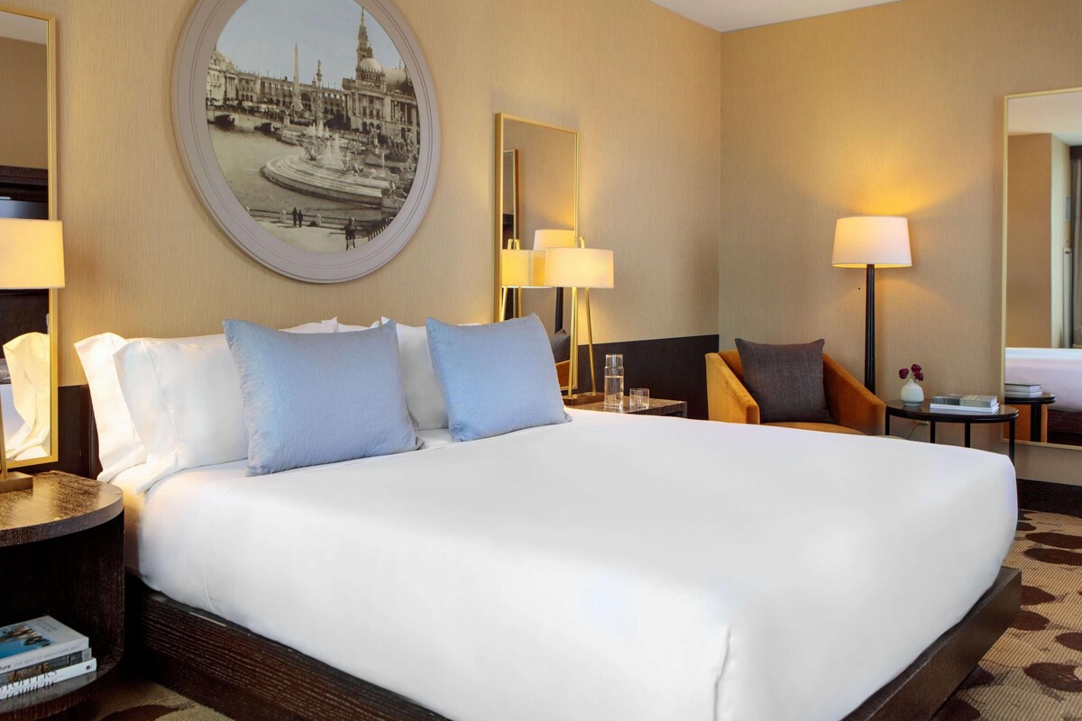 Een hotelkamer met een groot bed, twee nachtkastjes, een ronde spiegel en een aan de muur bevestigd schilderij.