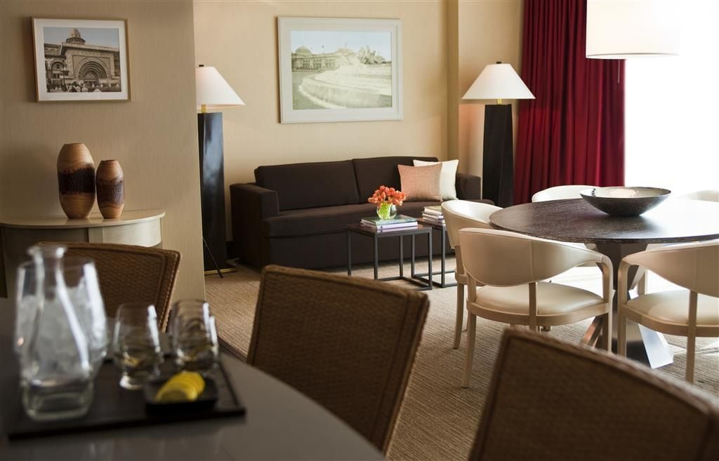 A hotel room with a dining area, a sofa, two lamps, and a table with a bowl and flowers.