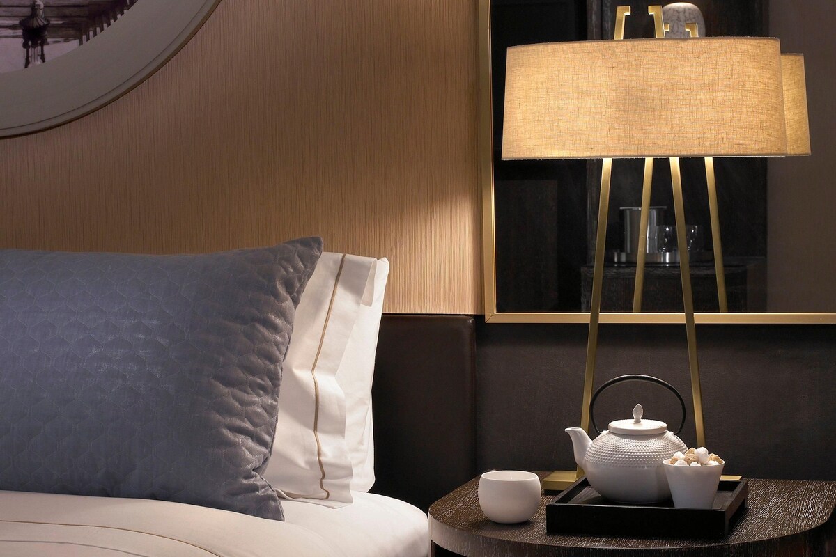 Een opgemaakt bed met kussens, een nachtkastje met een lamp, een theepot en een dienblad met kopjes.