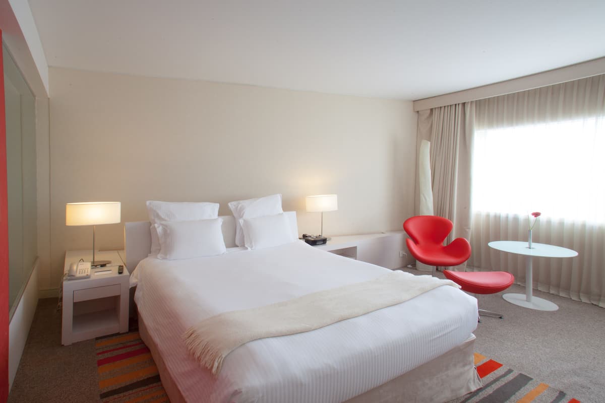 Habitación de hotel con una cama grande, dos mesitas de noche con lámparas, una silla roja, una mesita redonda y una ventana con cortinas.