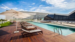 Zona de piscina con sillones de playa y sombrillas, un edificio moderno con amplios ventanales y un cielo azul despejado.