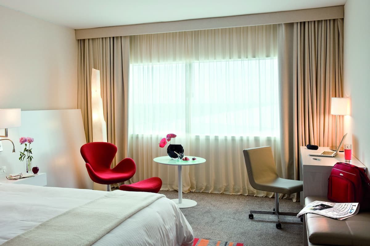 Habitación de hotel con cama, dos sillas, una mesita, lámpara y ventana con cortinas.