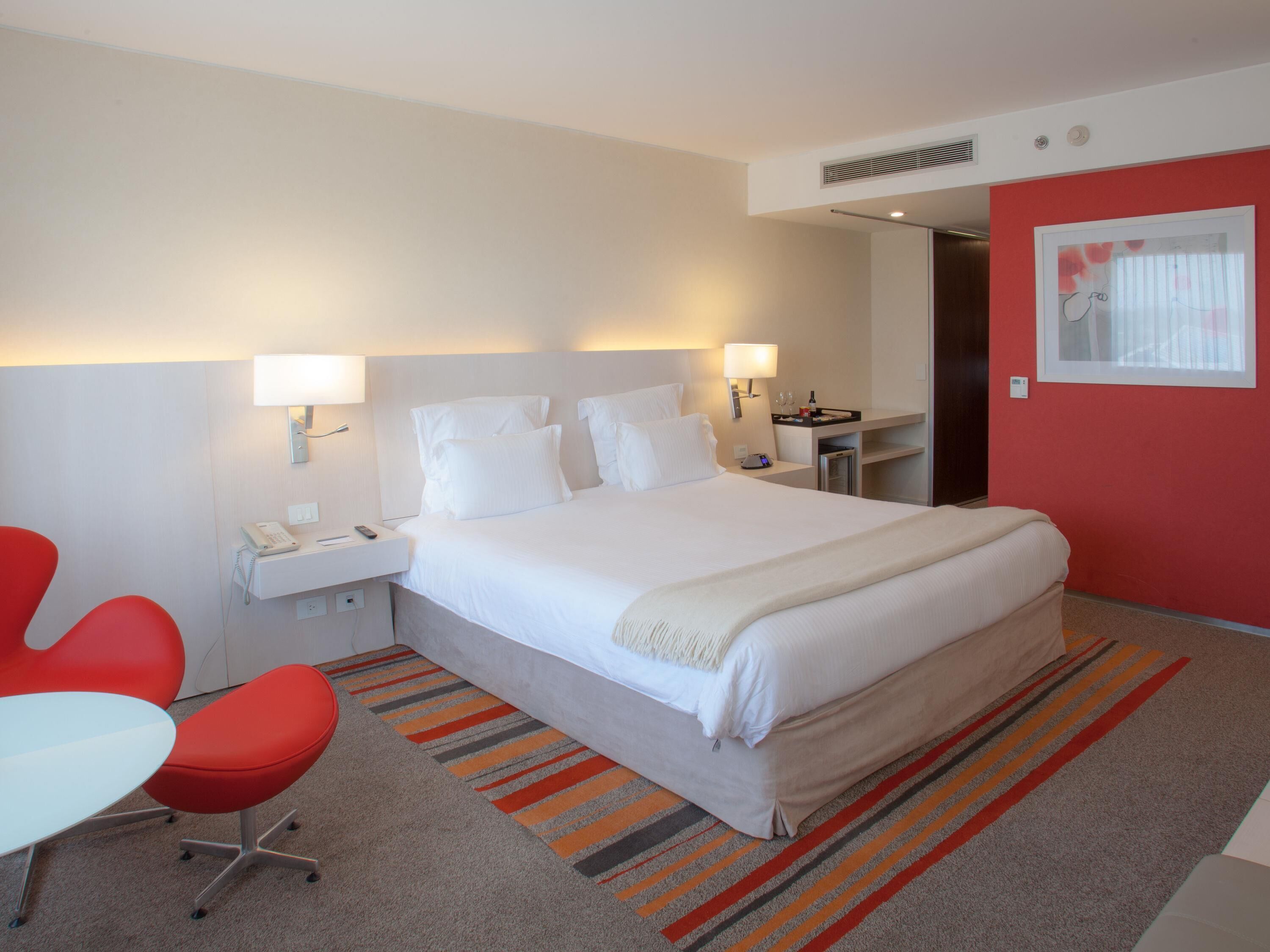 A hotel room with a large bed, two bedside lamps, a desk with a phone, a red chair, and a striped carpet.
