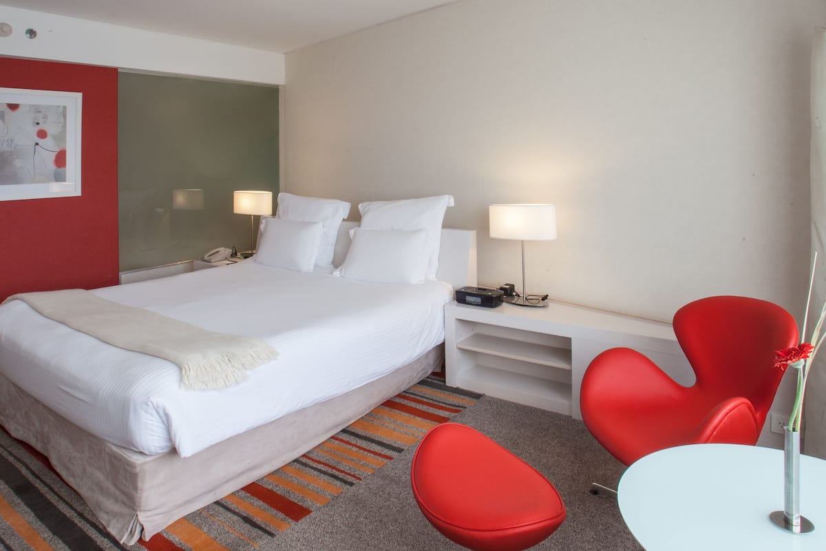 Habitación de hotel con una cama grande, dos lámparas de noche, un teléfono, una mesita con una silla roja y un florero rojo.